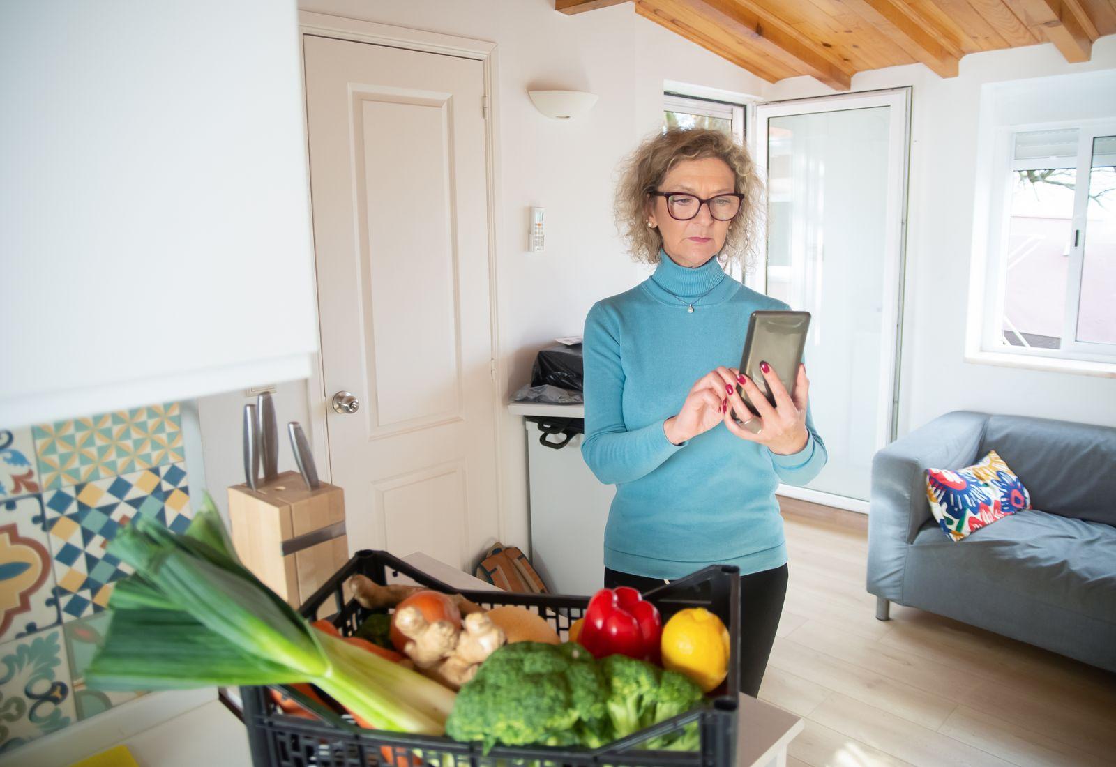 A woman checks her phone in her kitchen, next to a basket of groceries