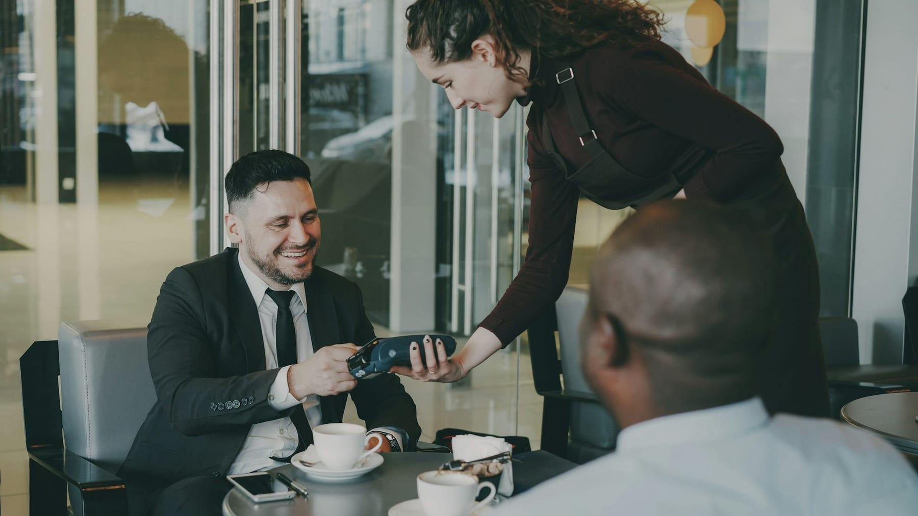 A diner pays by phone at a café table while the server holds the terminal
